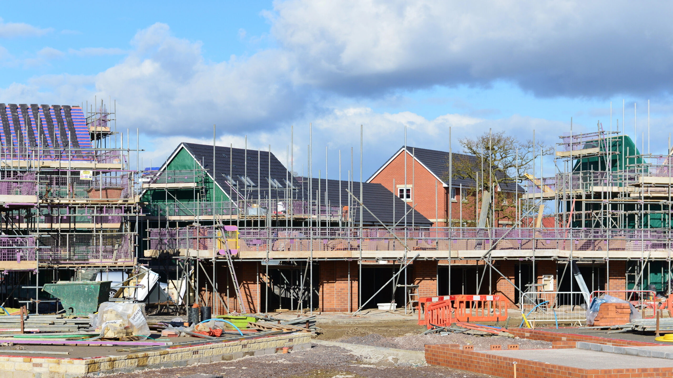 New houses under construction with scaffolding, building materials, and equipment scattered around the site. Some homes have roofs and windows installed, while others are still being built. Blue sky with clouds in the background.