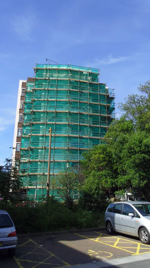 A tall building covered in green scaffolding is surrounded by trees and parked cars in a parking lot. The sky above is clear and blue.