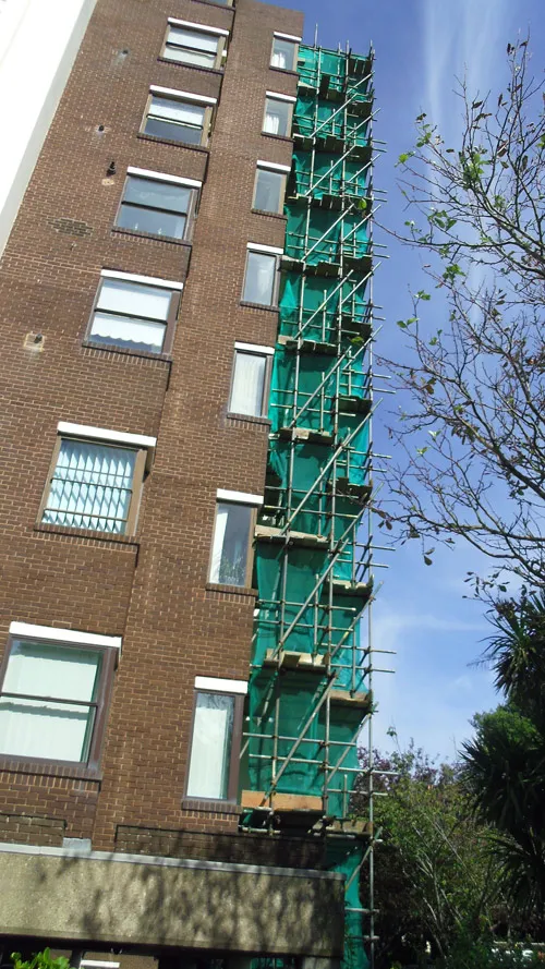 A multi-story brick building has scaffolding with green netting covering one side. There are windows on each floor visible on both the building and the scaffolding. Trees with green leaves are in the foreground under a clear blue sky.