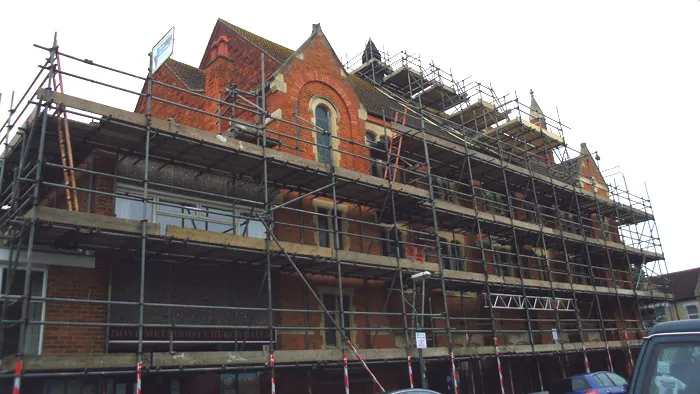 A large brick building with pointed roofs is surrounded by metal scaffolding. The scaffolding spans multiple stories, indicating ongoing construction or renovation work. Several cars are parked in front of the building. The sky is overcast.