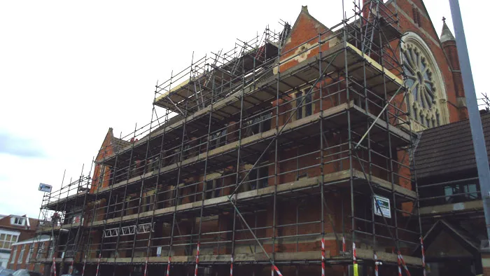 A building is under renovation with multiple layers of scaffolding covering its facade. The structure has a large circular stained glass window and pointed arches, indicative of Gothic architecture. Nearby smaller buildings and a cloudy sky can be seen.