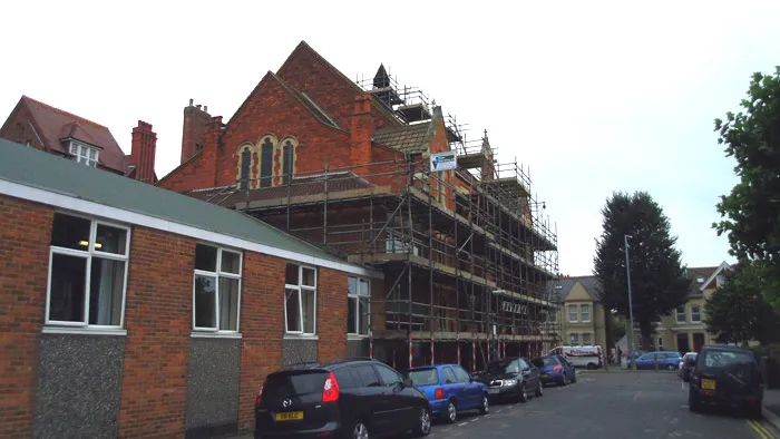 A brick building under renovation is covered in scaffolding. Several cars are parked along the roadside near the building. Another red brick building is visible on the left, and more residential buildings can be seen along the street.