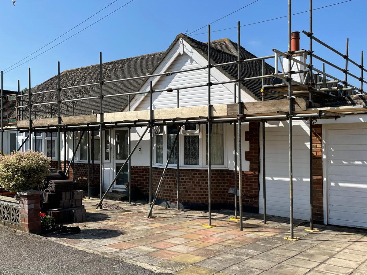 A one-story house with scaffolding set up along the front side, indicating ongoing construction or renovation work. The house has a mix of brick and white siding with a tiled roof, and the area in front features a paved driveway and some plants.