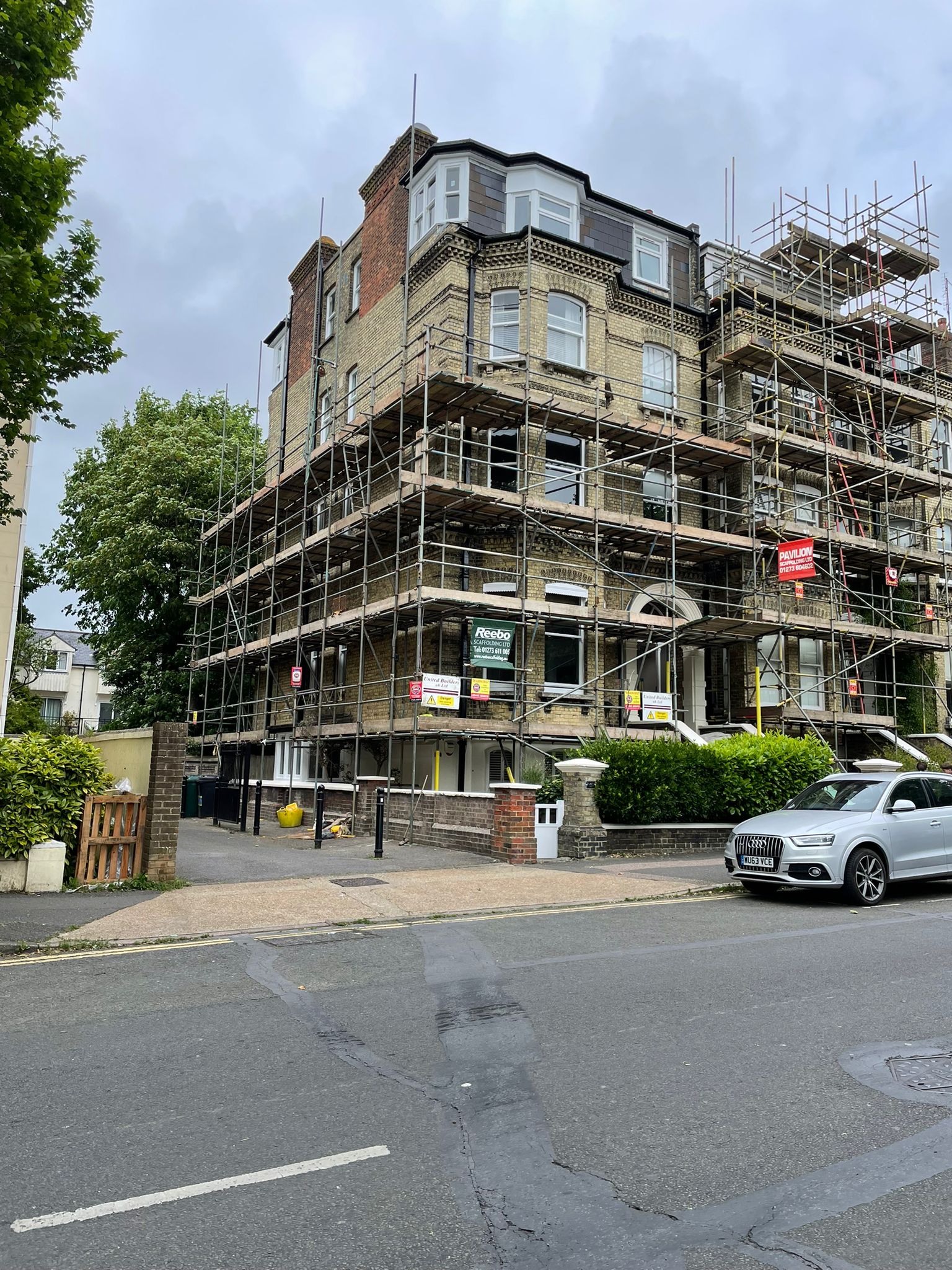 A multi-story building under renovation with extensive scaffolding covering its facade. The building is constructed with beige bricks and has large windows. A sign indicating construction is visible. A white car is parked on the street in front of the building.