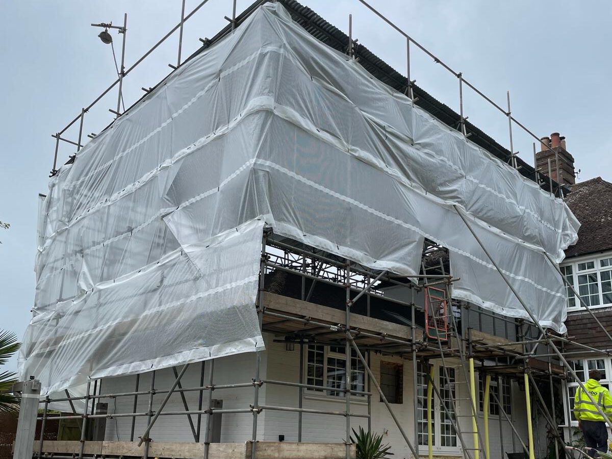 A house under renovation is covered in scaffolding and white protective sheeting. A worker in a high-visibility jacket stands on the right side. There are steps leading to a partially covered patio below, and a circular fire pit is visible in the foreground.