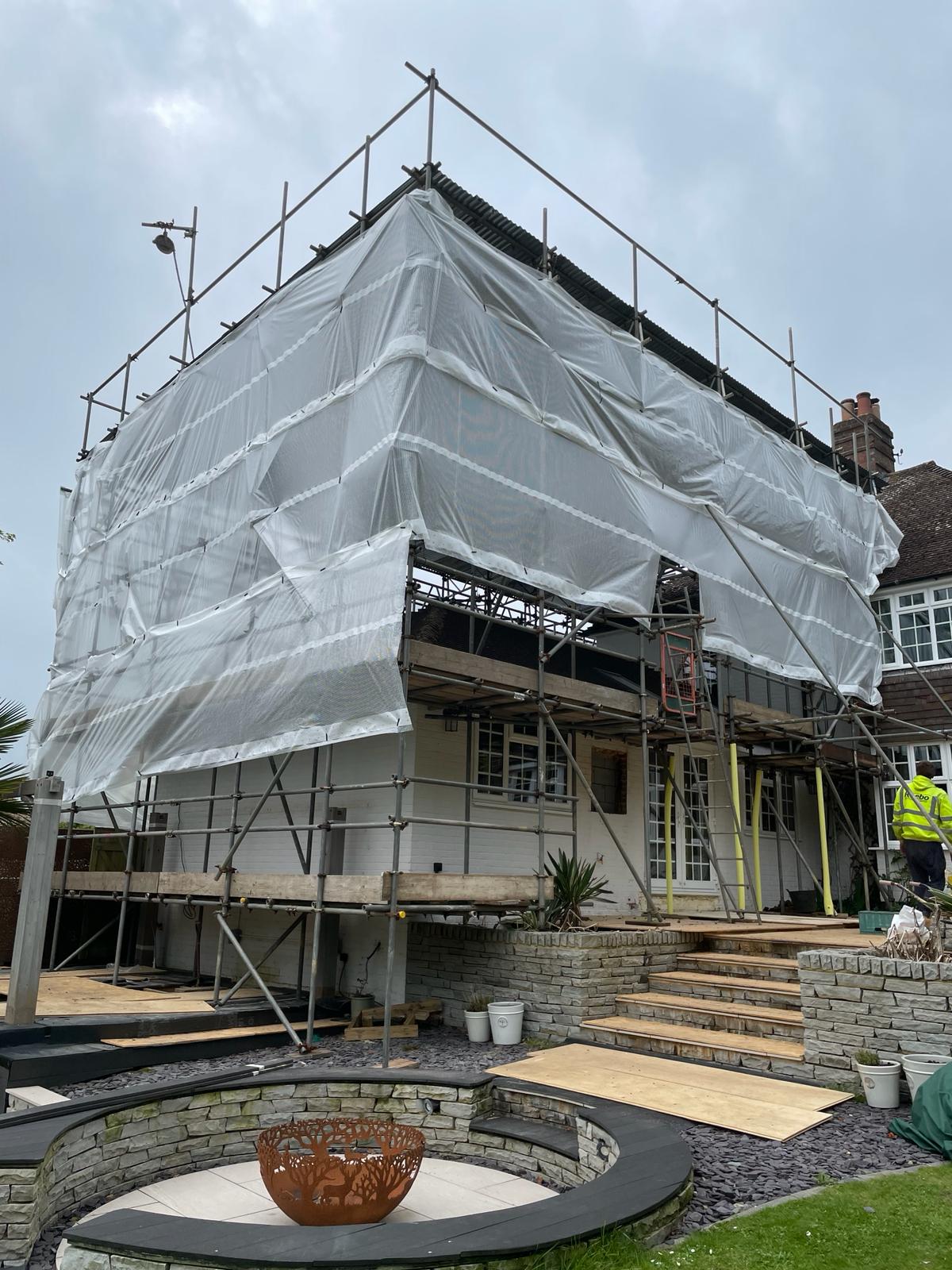 A house under renovation is covered in scaffolding and white protective sheeting. A worker in a high-visibility jacket stands on the right side. There are steps leading to a partially covered patio below, and a circular fire pit is visible in the foreground.