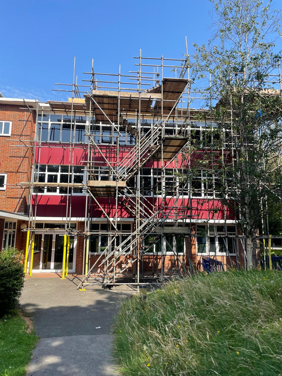 A three-story building under construction with scaffolding covering its facade. The building has a mix of red, beige, and brick exterior walls. Part of the building's surroundings includes a grassy area and trees, with a clear blue sky overhead.