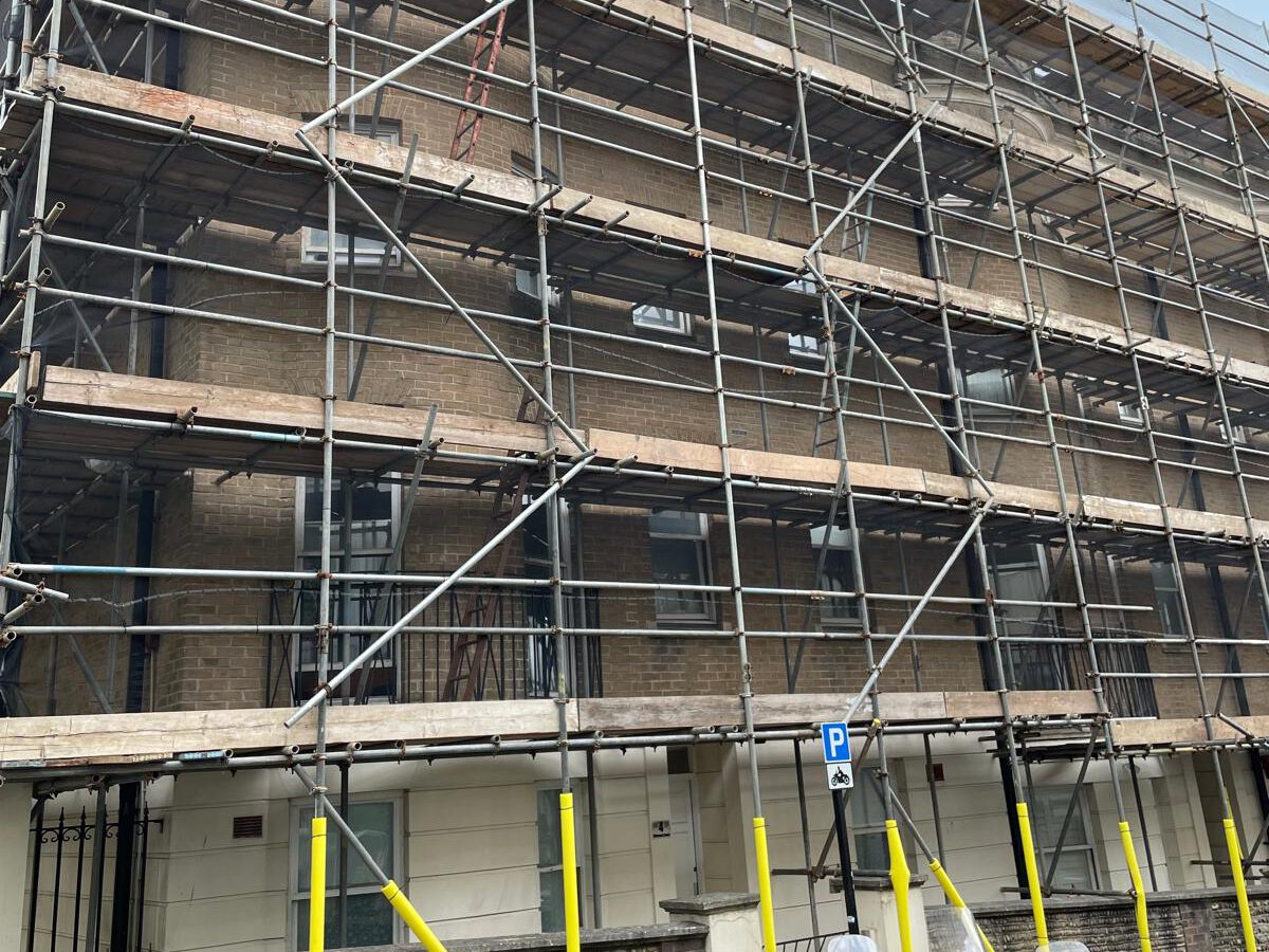 A multistory brick building is covered in scaffolding for construction or renovation. The scaffolding is made of metal bars and wooden planks. There's a blue parking sign with a white "P" on the street near the building.