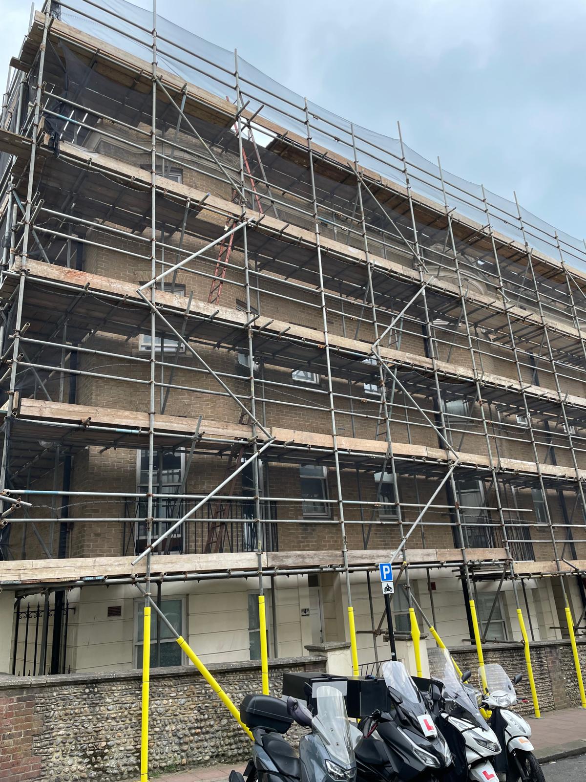 A multi-story building under renovation is covered in scaffolding. Several parked scooters, some with protective covers, are lined up in front of the building. Yellow safety barriers are positioned near the scaffolding, and the sky is overcast.
