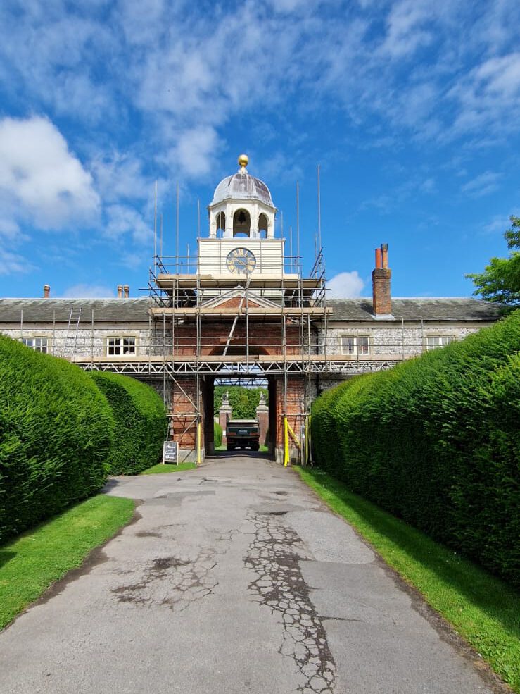 A pathway lined with tall green hedges leads to an archway with scaffolding. The archway features a clock and a dome on top. The sky is bright blue with scattered white clouds.