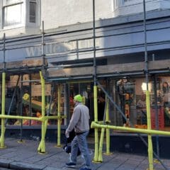 A storefront covered in scaffolding and yellow safety barriers on a street. The shop has large windows displaying various items. A person in a bright cap walks past, and another person in jeans and a blue jacket is visible in the background.