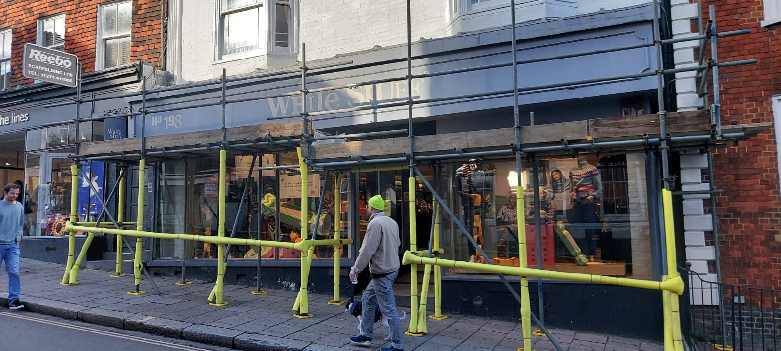 A storefront covered in scaffolding and yellow safety barriers on a street. The shop has large windows displaying various items. A person in a bright cap walks past, and another person in jeans and a blue jacket is visible in the background.