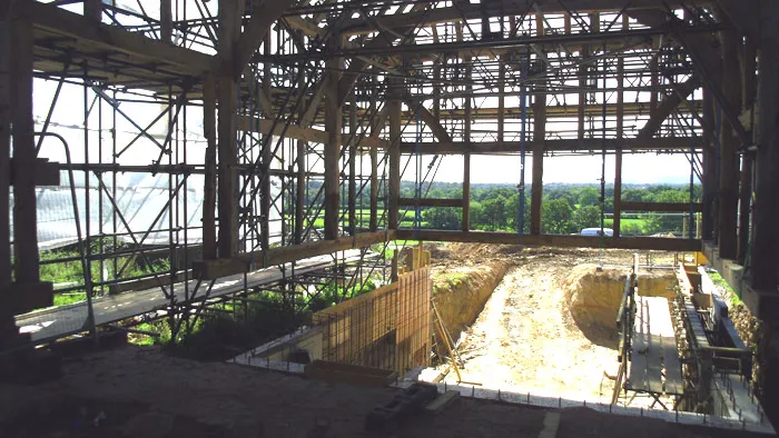 An unfinished construction site with exposed beams and scaffolding. The structure is partially built around a large pit or foundation area. The background shows a landscape with greenery and trees, indicating the location is in a rural or suburban area.