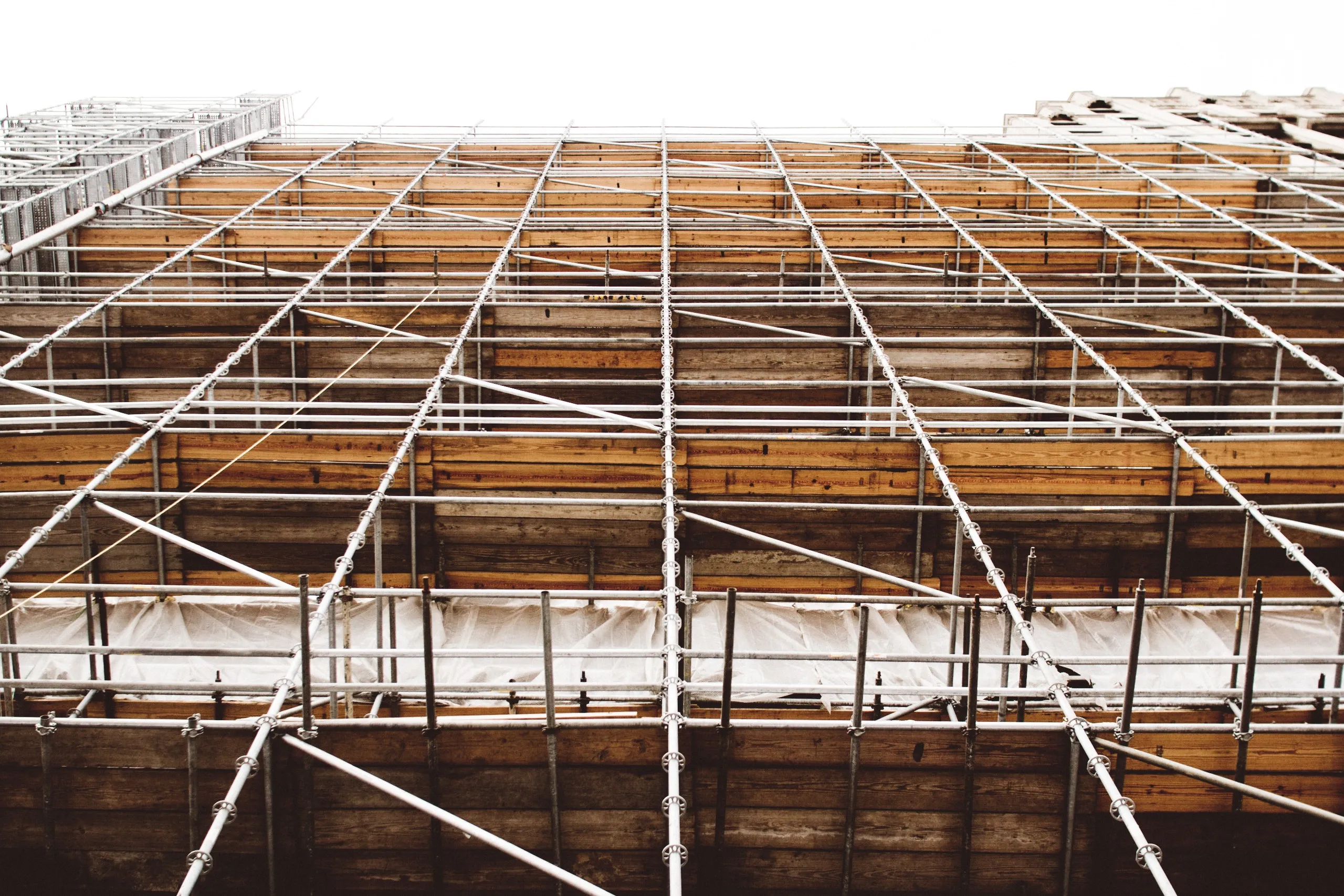 A low angle view of scaffolding on a building under construction. The metal framework and wooden planks create a grid-like pattern against a bright sky. The structure appears intricate and symmetrical, indicating a large and complex construction project.