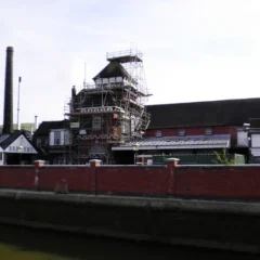 A large industrial building with a tall chimney and scaffolding around part of it. The building has several pitched roofs and a sign reading "Harvey's Brewery" is visible. A red brick wall runs along the front, bordering a river or canal.