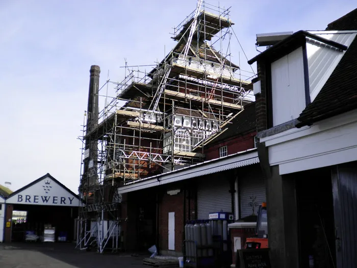 A brewery undergoing restoration work, with scaffolding surrounding its tall, multi-level brick structure. A nearby building with a white and brown exterior is visible, and an entrance sign to the brewery is seen on the left. The sky is clear and blue.