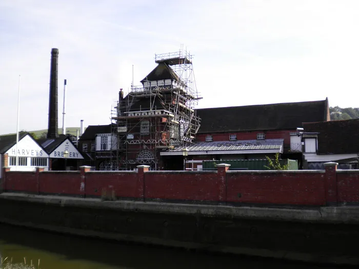 A large industrial building with a tall chimney and scaffolding around part of it. The building has several pitched roofs and a sign reading "Harvey's Brewery" is visible. A red brick wall runs along the front, bordering a river or canal.