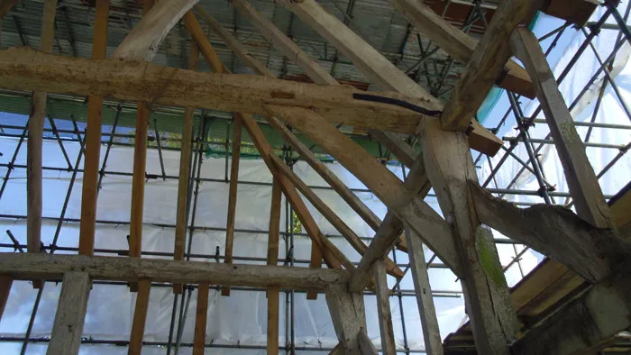 A view of a partially constructed or renovated building with wooden beams and scaffolding. The structure is surrounded by a transparent covering and metal framework, allowing natural light to filter through. Tools and construction materials are visible in the background.