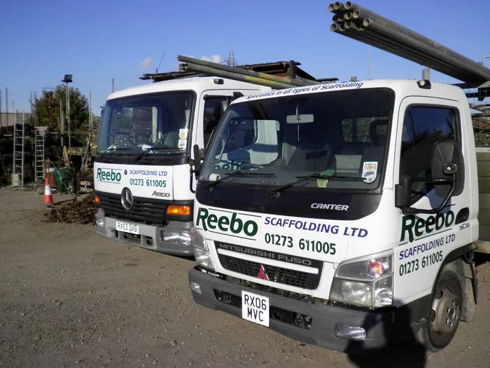 Two white scaffolding trucks with the text "Reebo Scaffolding LTD" and a contact number on the front parked side by side in an outdoor storage yard. The trucks are loaded with long metal scaffolding pipes, and various construction materials are visible in the background.