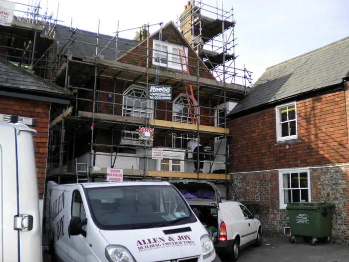 A multi-story brick house is under extensive renovation with scaffolding on the front facade. Signs are visible on the scaffolding. Vans, including one labeled "Allen & Joy Builders Contractors," are parked in front. A green dumpster is on the right.