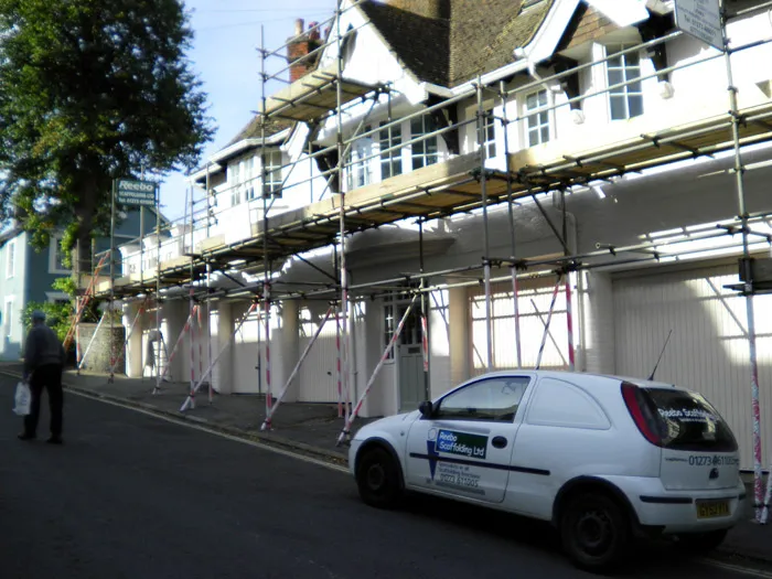 A white car with "Apollo Scaffolding Ltd" signage is parked on a street. Large scaffolding is set up along the front of a white building with gabled roofs. A person carrying a bag walks on the sidewalk near the building. Trees are visible in the background.