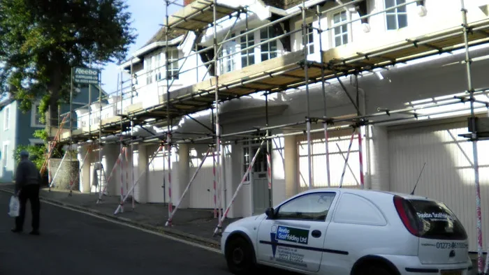 A row of terraced houses under renovation, with scaffolding erected in front. A man walks on the street carrying a plastic bag, and a small white car with "Rivals Scaffolding Ltd" branding is parked nearby. Trees and other buildings are visible in the background.