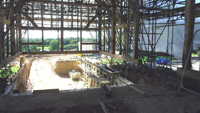 A large construction site inside a partially built structure with scaffolding. The site includes a deep rectangular excavation with building materials and equipment around. Sunlight streams in through the open sides, revealing a green field in the background.