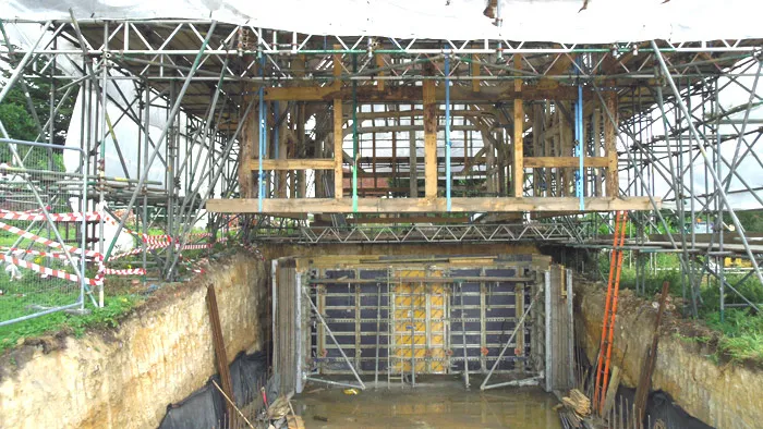 Construction site with scaffolding surrounding a structure that appears to be an entrance or portal. The area is excavated, with a noticeable trench and muddy ground. Metal framework and wooden planks support the structure, secured with caution tape around the site.