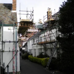 A narrow alley between white buildings filled with scaffolding on both sides, supporting ongoing construction and repair work. Some windows and doors are visible amidst the structure, and a narrow path runs through the middle, bordered by small shrubs and trees.