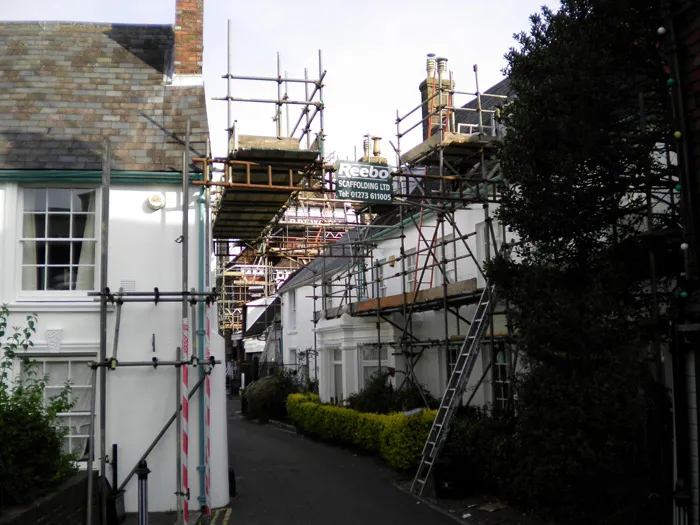 A narrow alley between white buildings filled with scaffolding on both sides, supporting ongoing construction and repair work. Some windows and doors are visible amidst the structure, and a narrow path runs through the middle, bordered by small shrubs and trees.