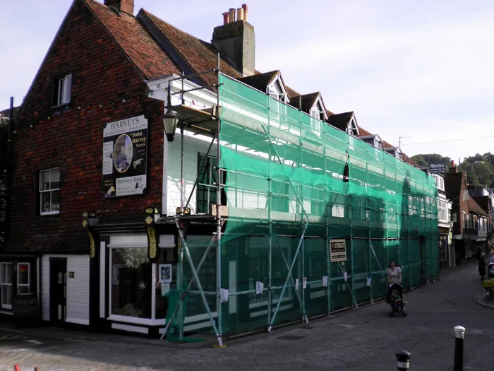 A two-story building with a steep roof under renovation is covered in green scaffolding on a street corner. Signs for "Harveys" and other businesses are visible. A person pushing a stroller walks past on the sidewalk below. The weather appears to be clear and sunny.