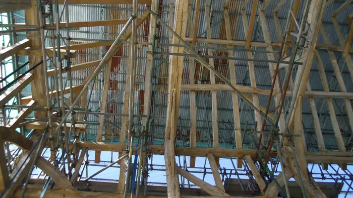 A view from below of a construction site showcasing a complex framework of wooden beams and metal scaffolding. The structure extends upward, with sunlight filtering through gaps in the partially completed roof.