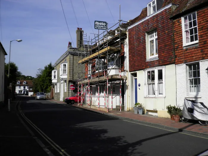 A narrow residential street with scaffolding set up on a two-story house for repair or renovation. The house is painted white with red-tiled upper walls. Several potted plants sit on the brick sidewalk. Other similar houses line the street, which is quiet and empty.