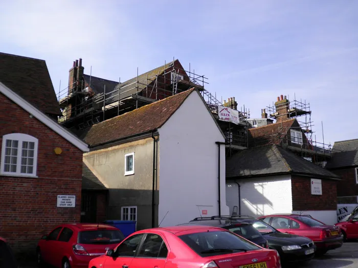 A row of old brick and plaster buildings under renovation, with scaffolding set up on the roofs and walls. Several red and gray cars are parked in the foreground. Signs on the construction site provide safety information. The sky is clear and sunny.