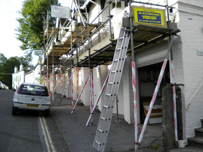 A row of white buildings is under construction with scaffolding in place. A tall metal ladder leans against the scaffolding, and a small white car is parked nearby. Various signs are attached to the scaffolding, including one for an alarm system. 

.