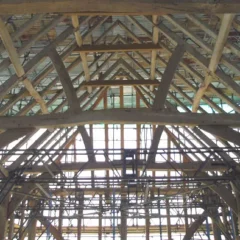 A view of a timber-framed building under construction. The image shows a complex framework of wooden beams and trusses forming the structure of the roof, with light filtering through gaps. Scaffolding can be seen supporting sections of the framework.