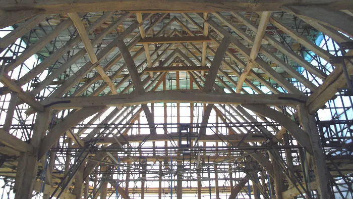 A view of a timber-framed building under construction. The image shows a complex framework of wooden beams and trusses forming the structure of the roof, with light filtering through gaps. Scaffolding can be seen supporting sections of the framework.