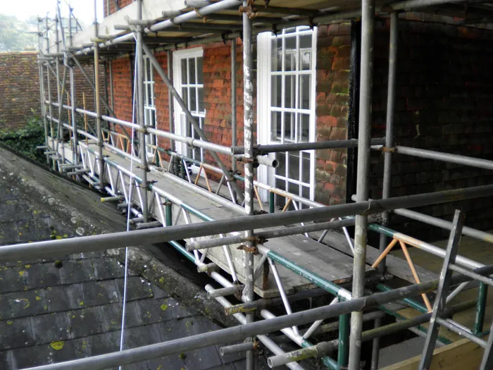 Scaffolding erected along the side of a brick building, providing access to windows and the exterior wall. The structure consists of metal pipes and wooden planks, positioned above a sloped roof with some debris visible on the surface.