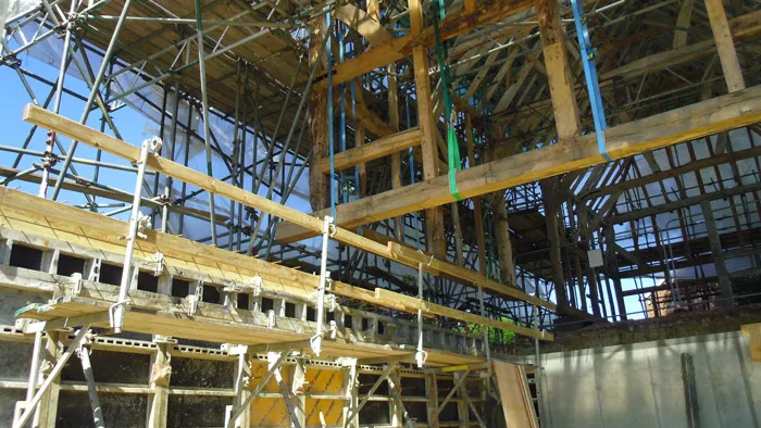 A construction site with a wooden framework in the foreground supported by metal scaffolding. Various beams and planks are visible, along with metal rods and braces. Sunlight illuminates parts of the structure, casting shadows. Blue sky in the background.