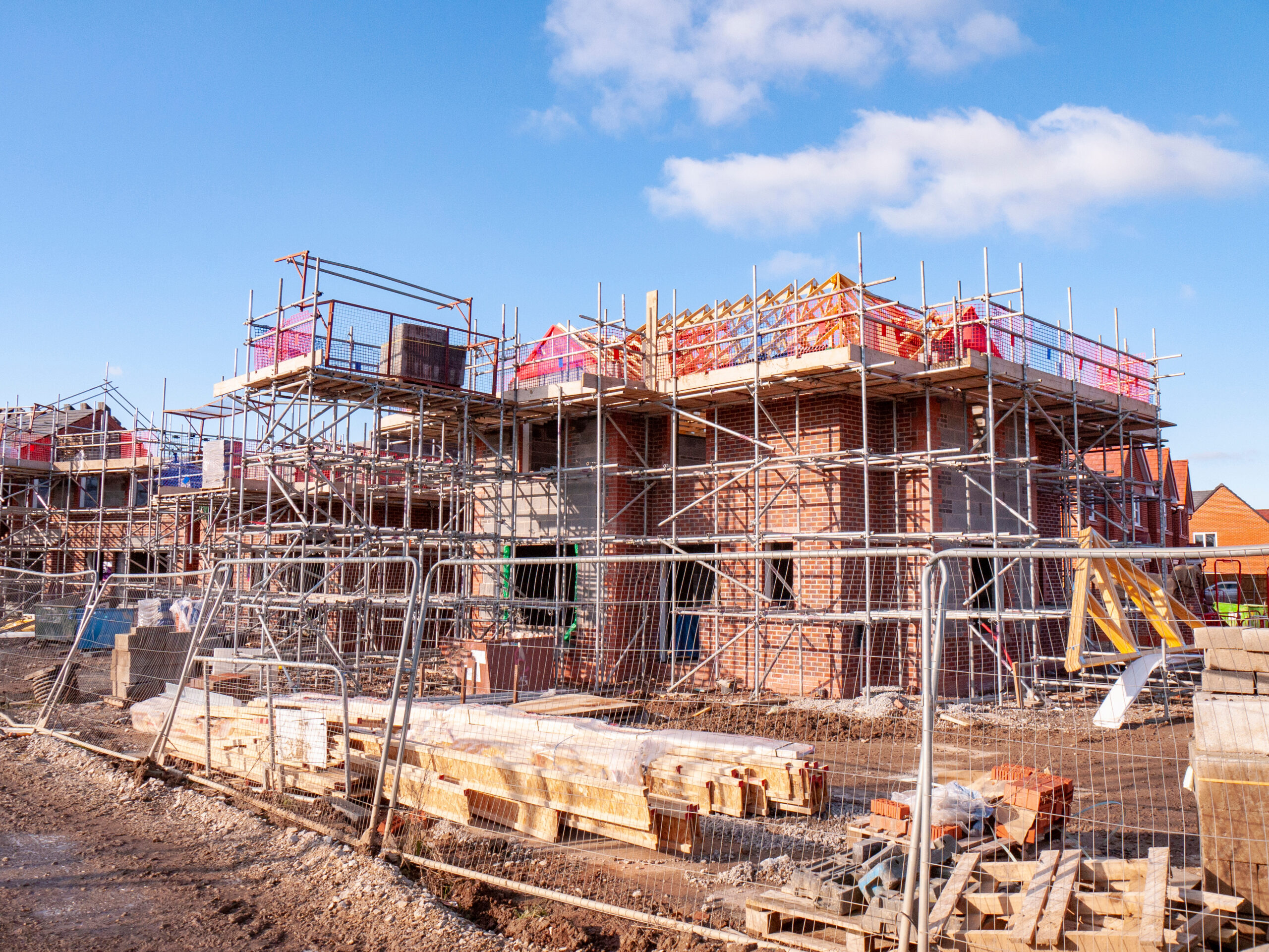 A construction site with partially built brick houses, surrounded by scaffolding and safety barriers. Building materials and equipment are scattered around the area under a clear blue sky.