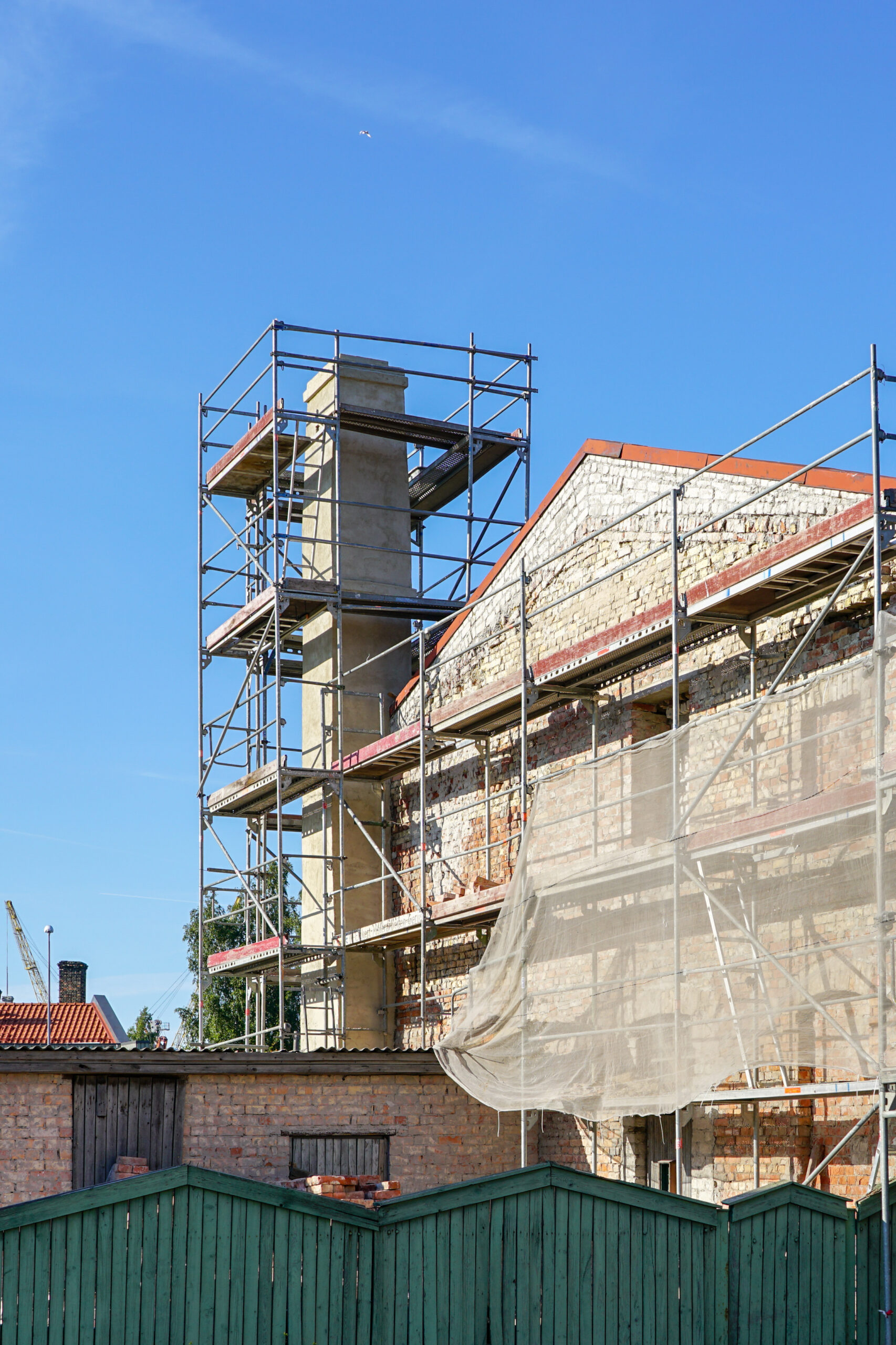 A building under renovation with scaffolding and netting covering its partially exposed brick walls, set against a clear blue sky. Green wooden fences are visible in the foreground.