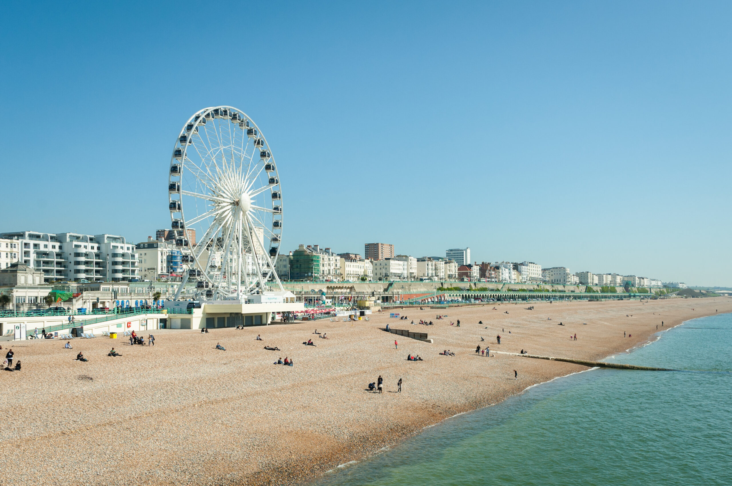 A large Ferris wheel stands on a pebbled beach with scattered people relaxing. Modern buildings and a clear blue sky line the background, and calm sea water is visible on the right.