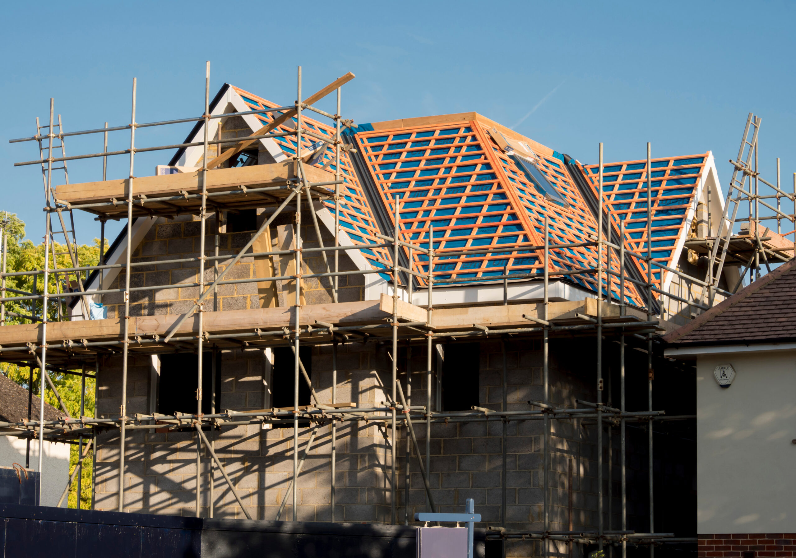 A partially constructed house with scaffolding around it, showing exposed brick walls and an unfinished roof with visible wooden beams and blue weatherproofing membrane under a clear blue sky.