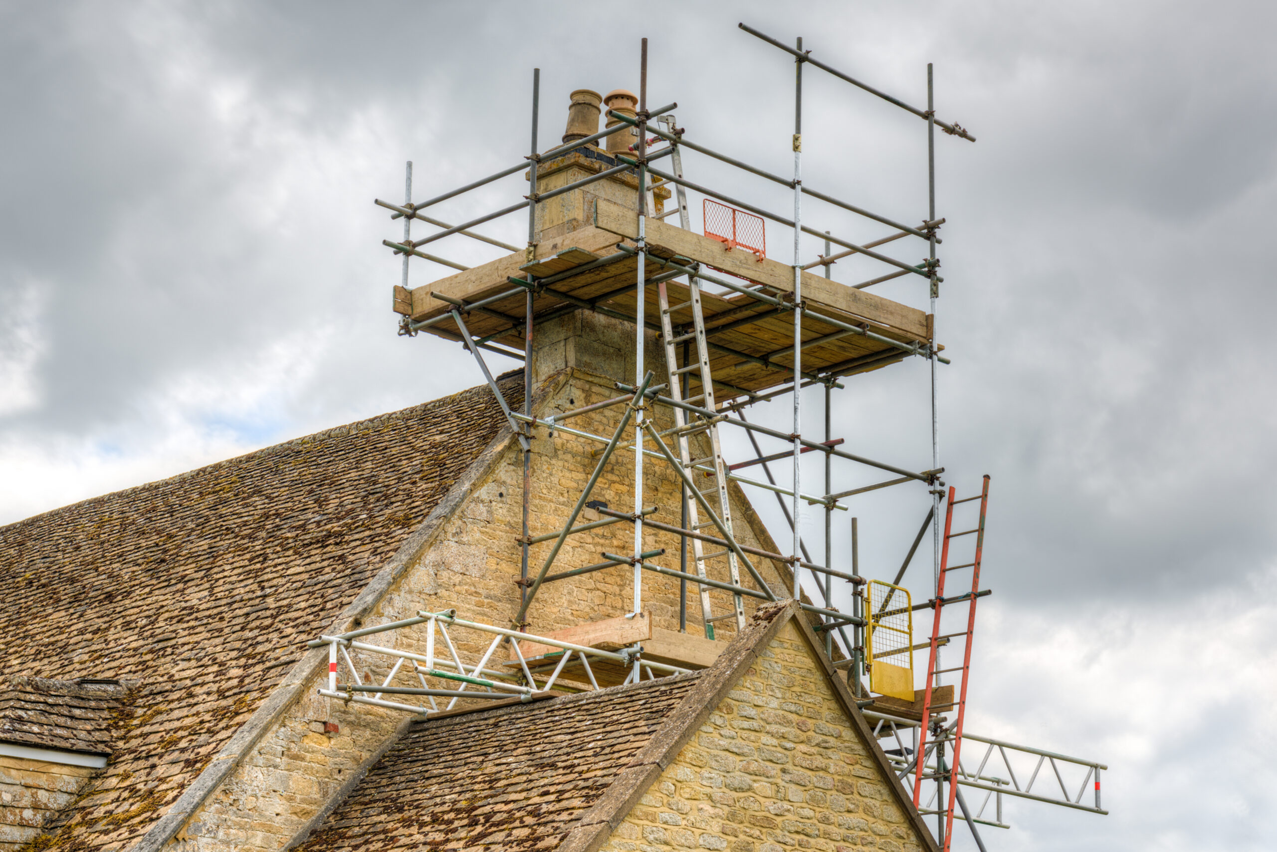 A stone house roof with scaffolding built around the chimney for repair or maintenance work, under a cloudy sky. Several ladders and metal platforms are attached to the scaffolding structure.