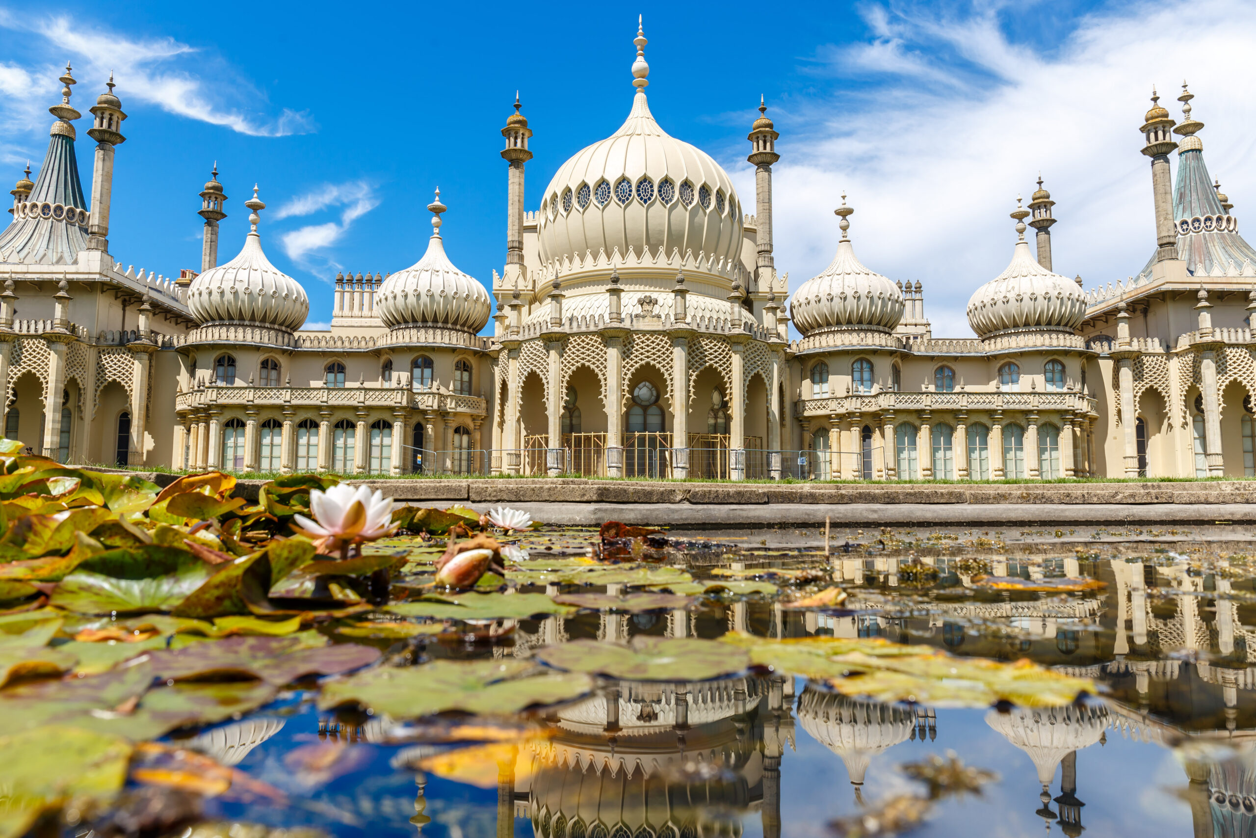 The Royal Pavilion in Brighton, England, with its ornate domes and minarets, is reflected in a pond filled with lily pads and water lilies under a bright blue sky with scattered clouds.