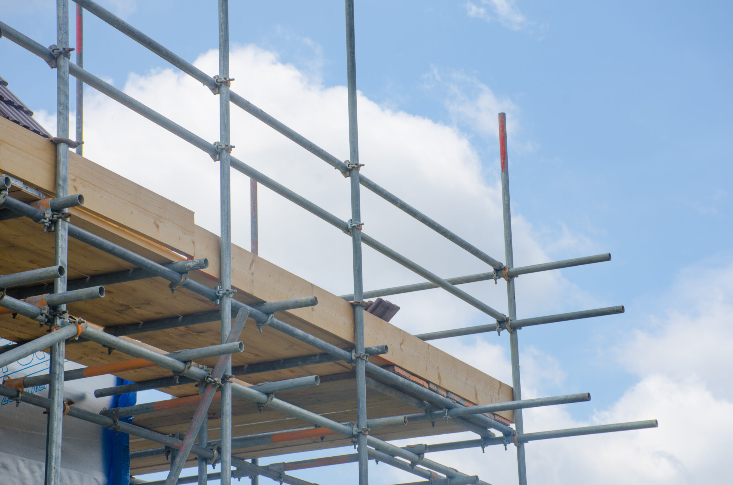 Scaffolding and wooden planks set up on the side of a building under construction, with a bright blue sky and scattered white clouds in the background.
