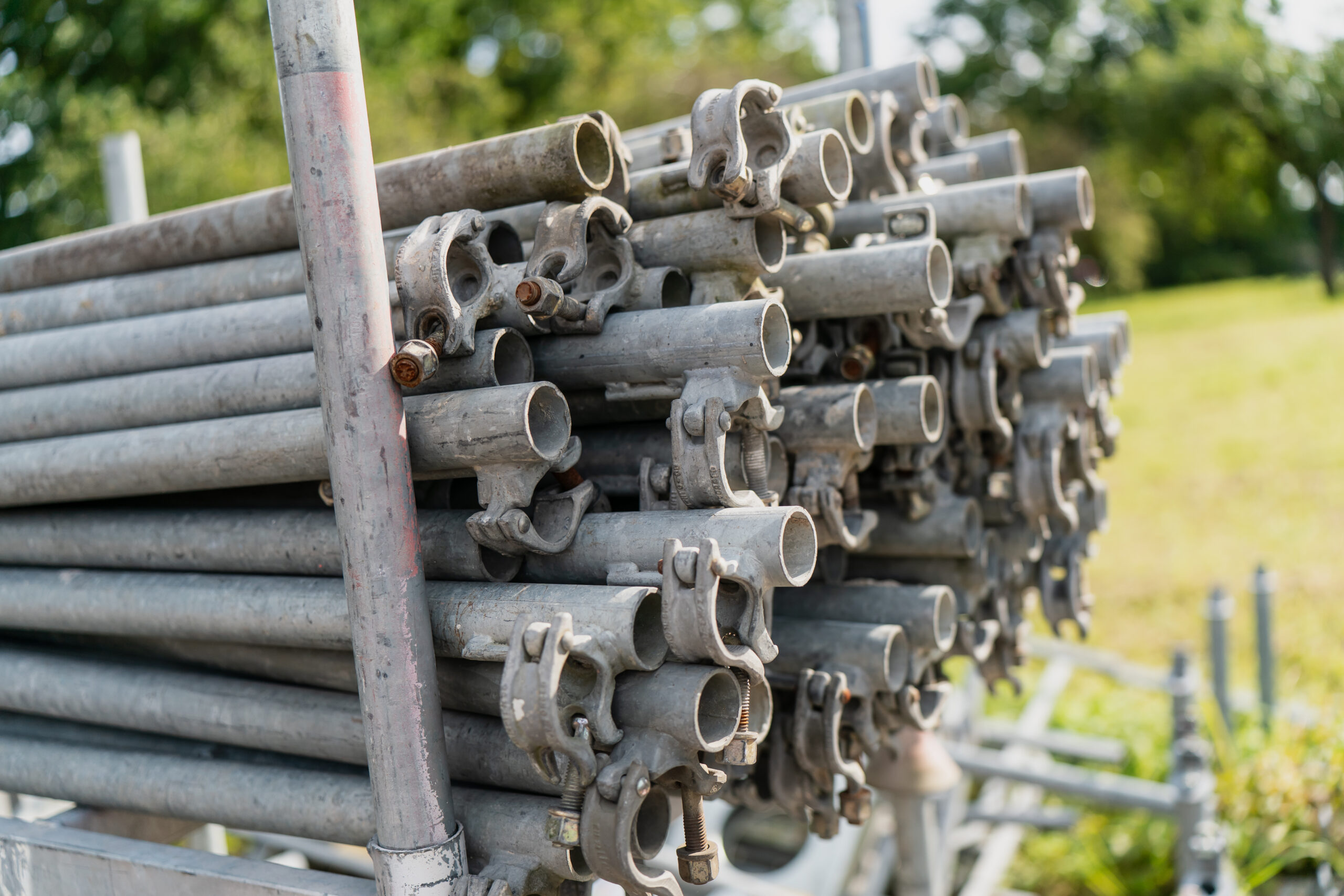 A stack of metal scaffolding pipes with clamps is grouped together outdoors on a sunny day. Green grass and trees are visible in the background.