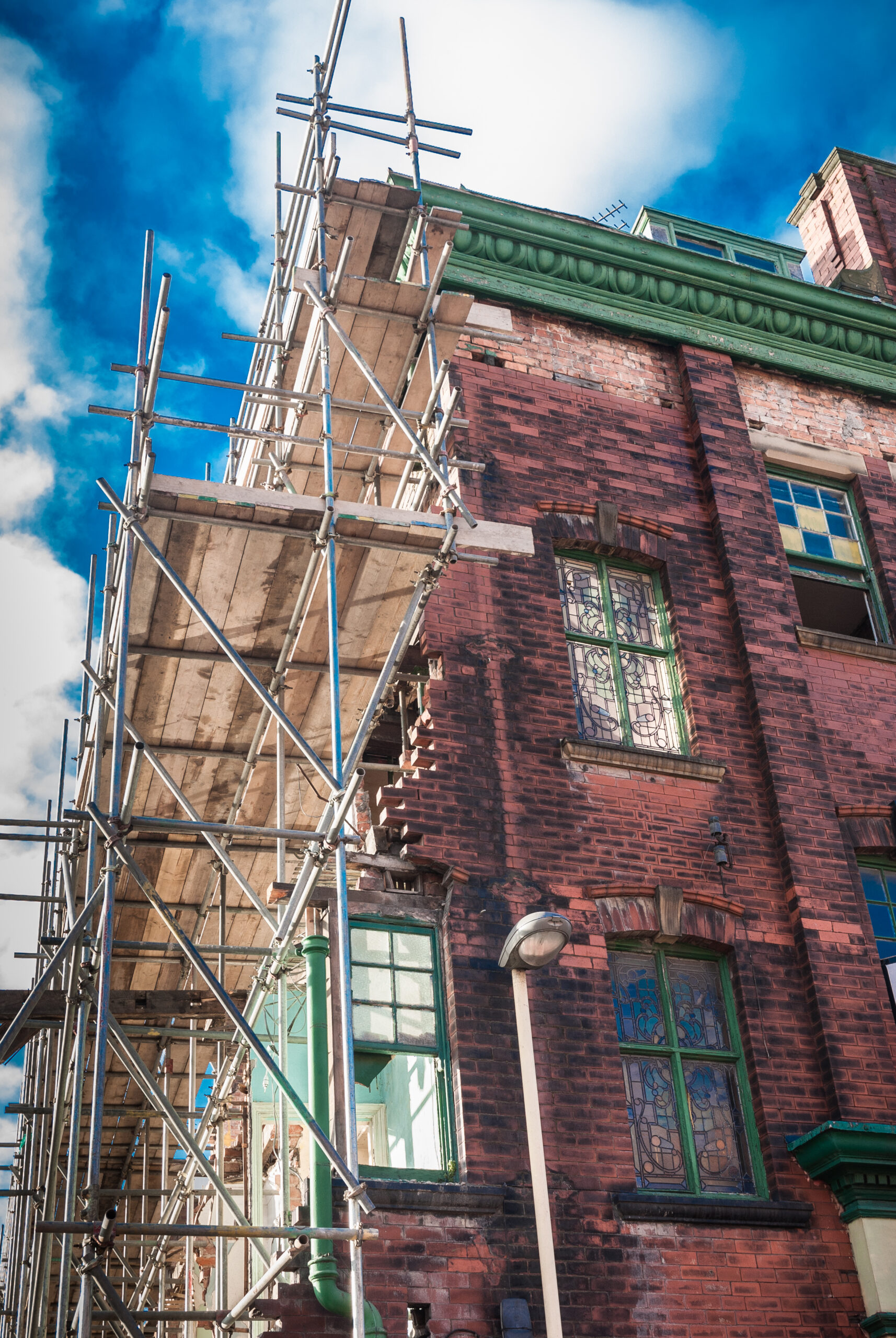 Scaffolding surrounds the upper floors of a red brick building with green-trimmed windows and peeling paint, under a partly cloudy blue sky.