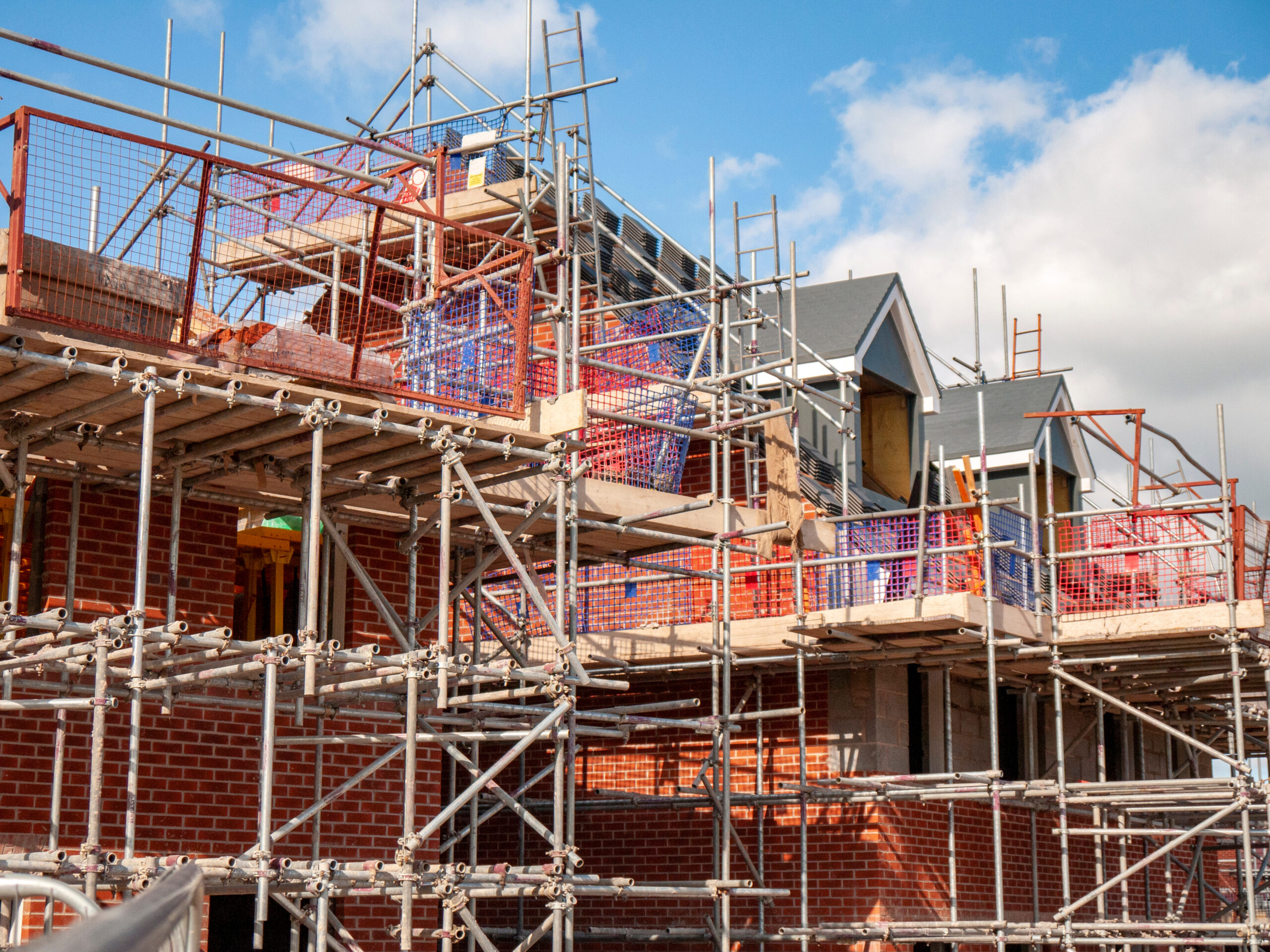 A partially constructed brick building surrounded by metal scaffolding under a blue sky with clouds. Some rooftops and windows are visible behind the scaffolding.