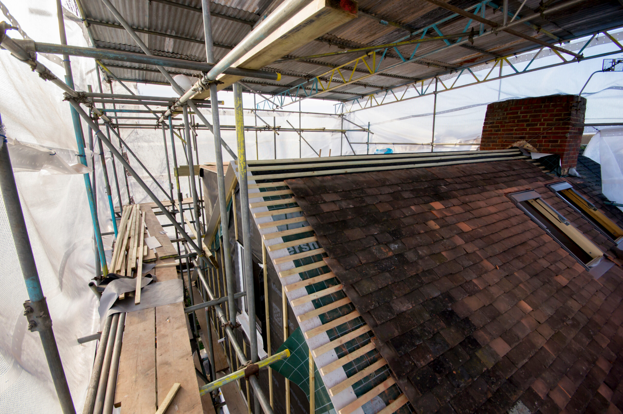 A partially tiled roof under construction, surrounded by scaffolding and covered with a temporary protective canopy; timber battens are visible where tiles have not yet been laid.